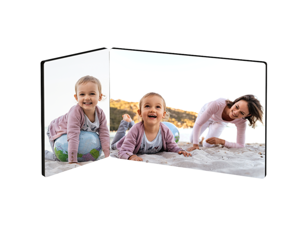 Ein zweiteiliger Fotoaufsteller zeigt eine fröhliche Szene am Strand. Auf der linken Seite sitzt ein kleines Mädchen auf einem bunten Ball und lächelt. Auf der rechten Seite krabbelt ein anderes kleines Mädchen im Sand und schaut lachend zur Kamera. Eine junge Frau kniet daneben im Sand und lächelt ebenfalls. Im Hintergrund ist ein heller Sandstrand und etwas Grün zu sehen.