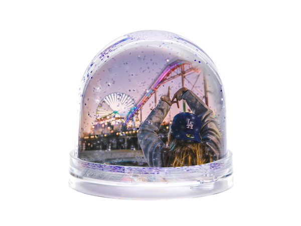 Glitzerkugel zeigt Mädchen mit Cap von hinten, formt mit Händen „LA“-Zeichen; im Hintergrund bunte Achterbahnen.