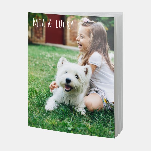 Ein fröhliches Mädchen sitzt lachend mit einem weißen Hund auf dem Rasen – Fotobuch A4 hoch für Tierliebe-Momente.